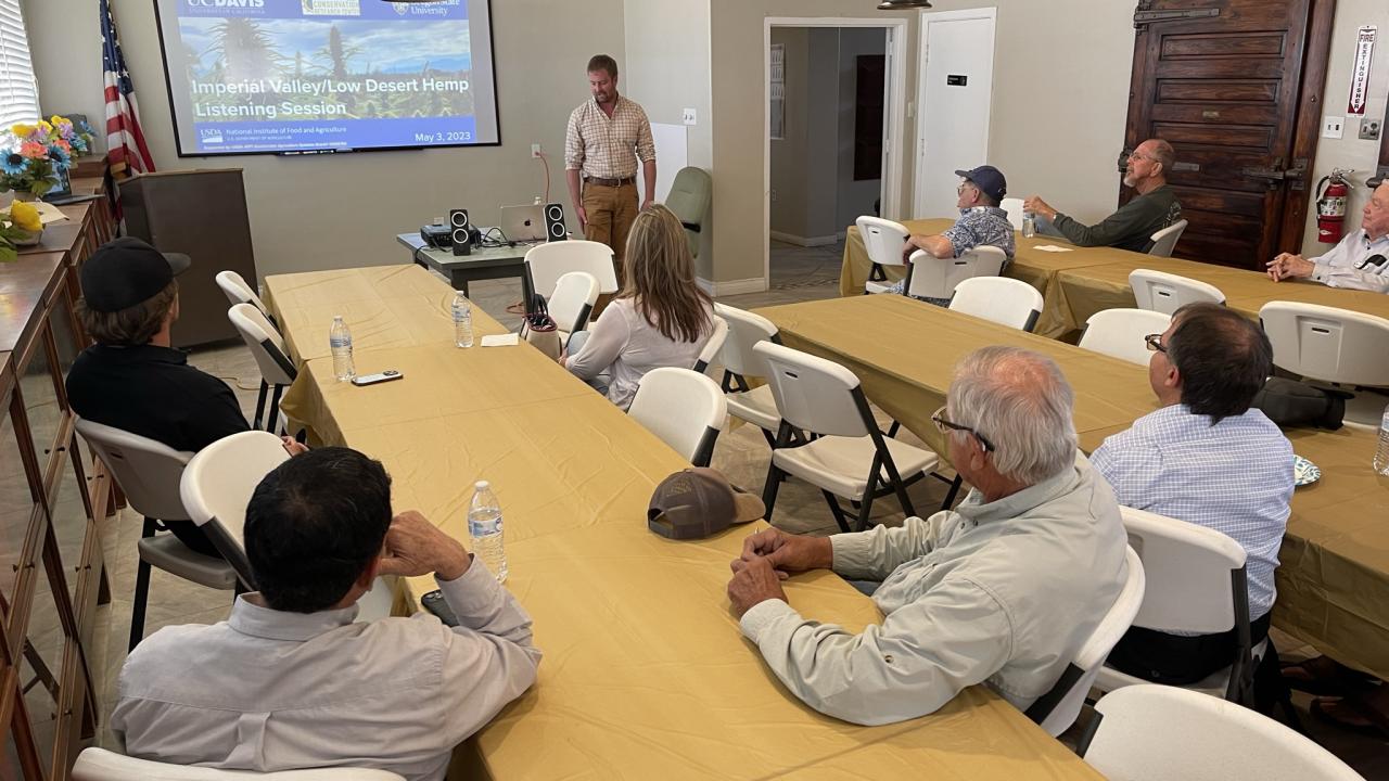 listeners sitting at a long wooden table watching a presentation 