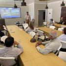 listeners sitting at a long wooden table watching a presentation 